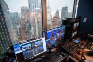The view through a window in a skyscraper in the NewYork looking across the skyline. In the foreground there is a livestream audio and video mixing desk.